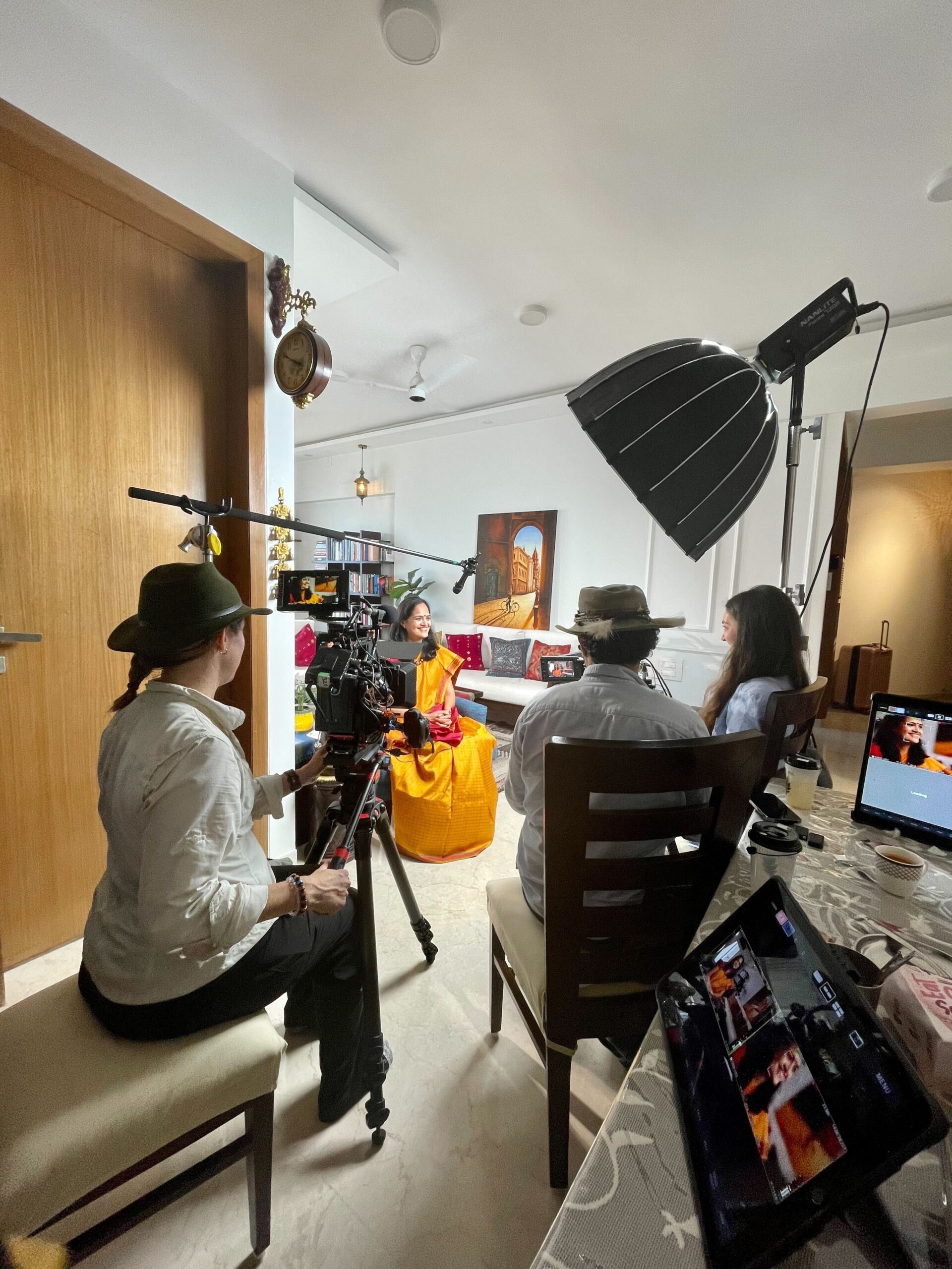A film crew records an interview in a well-lit living room. A woman in a bright yellow and orange sari sits on a white couch, smiling as she speaks. Around her, the crew operates cameras, lighting, and a boom mic, while screens and equipment are visible on a nearby table. The setting includes bookshelves, warm lighting, and a painting of an archway on the wall.